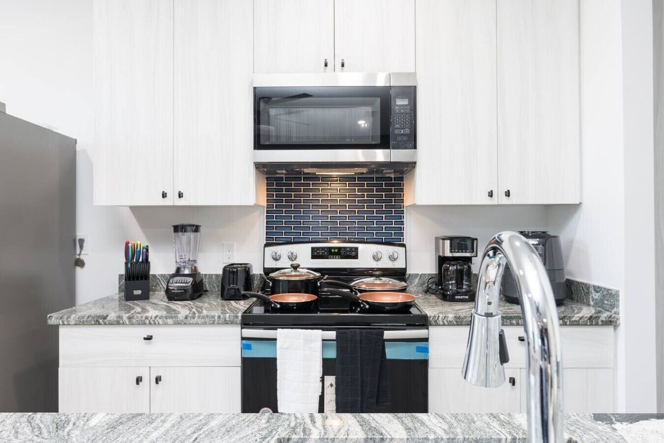 A modern kitchen with white cabinets, a black microwave, a stove with pots, a coffee maker, and a blender.
