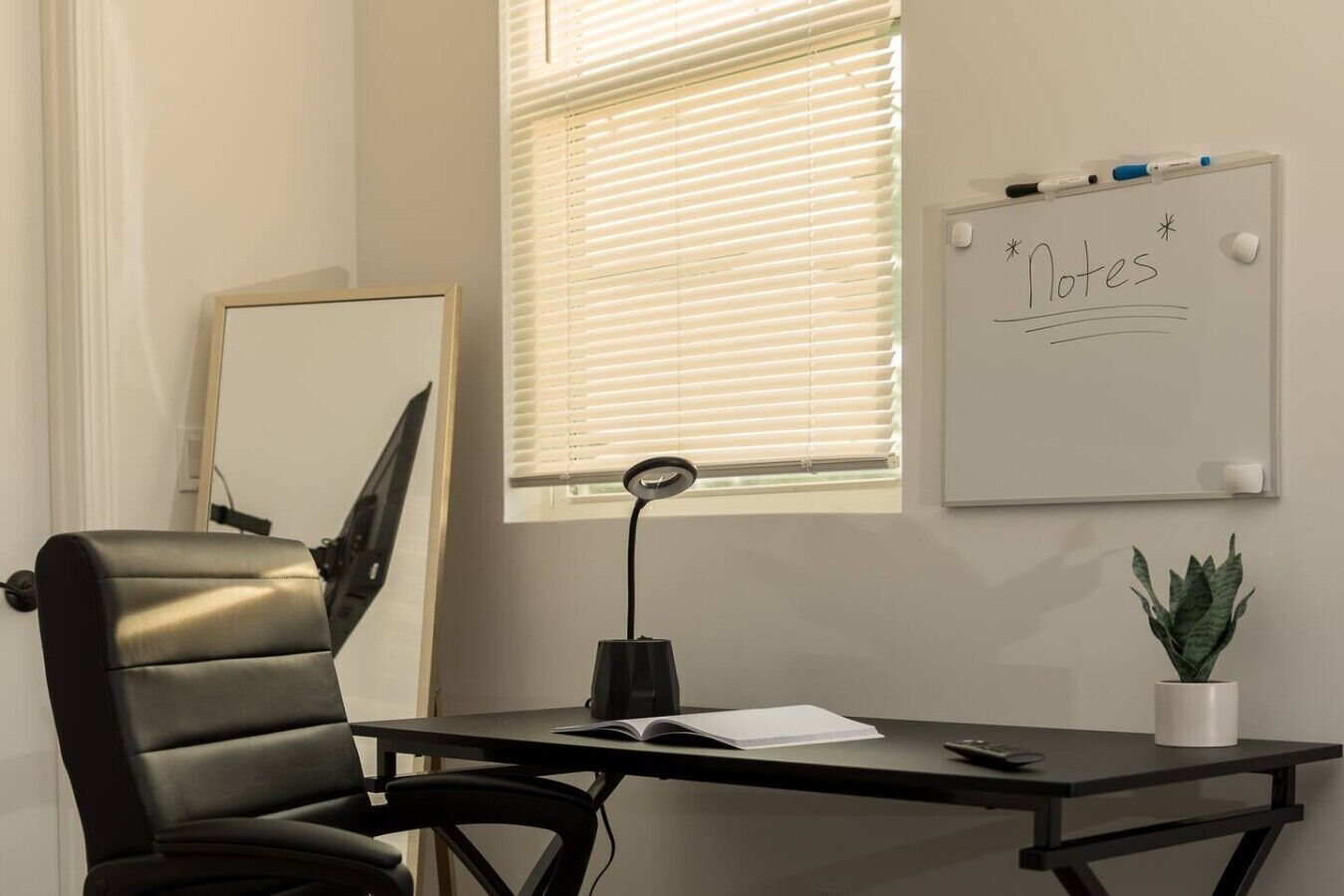 A conference room with a black leather chair, a desk with a laptop, a whiteboard with 'Notes' written on it, a window with blinds, and a potted plant.