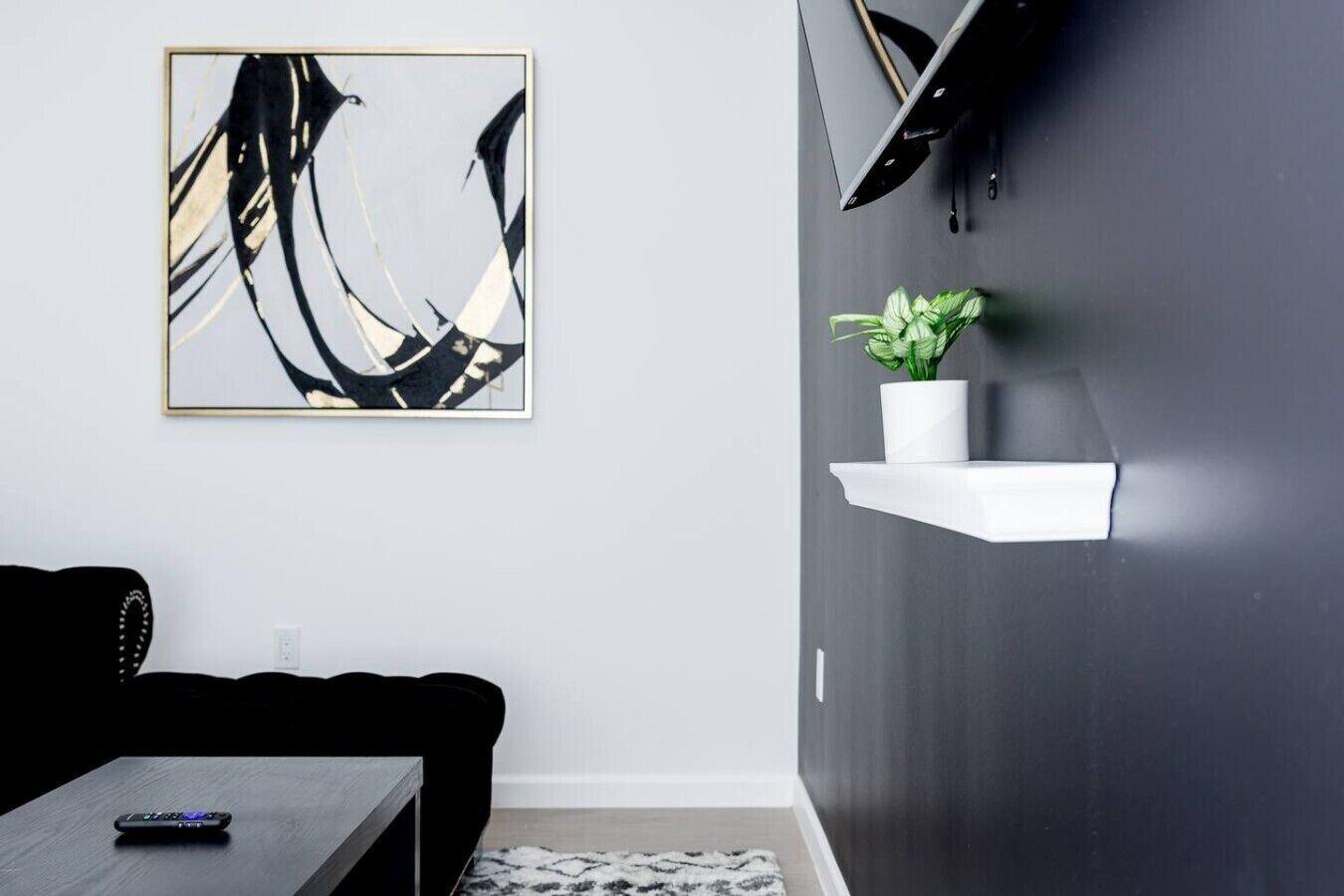 En moderne stue med en sort sofa, et sofabord, et indrammet abstrakt billede, et ophængt fjernsyn og en hvid reol med en plante.
