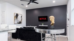Een moderne woonkamer met een zwarte bank, een witte salontafel, een televisie waarop 'NETFLIX' staat en een plafondventilator.