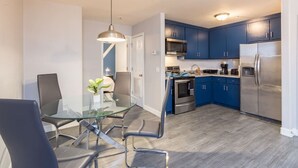 Een moderne keuken met blauwe kastjes, roestvrijstalen apparatuur en een glazen eettafel met stoelen.