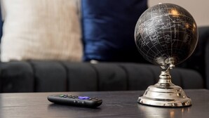 Een wereldbol op een metalen voet, een afstandsbediening, en een donkerbruine bank met kussens op de achtergrond.