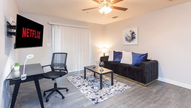A modern living room with a ceiling fan, a flat-screen TV, a black leather sofa, a coffee table, and a desk with a chair. - King Bed, Central Location  Fast WiFi / Long Term (McAllen)