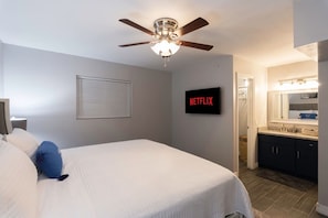 Makuuhuoneessa on sänky, kattotuuletin, seinälle asennettu televisio, jossa näkyy Netflix, sekä kylpyhuone, jossa on peili ja pesuallas.