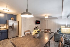A modern kitchen with a stainless steel refrigerator, a dining table with chairs, a TV mounted on the wall, and a ceiling fan.