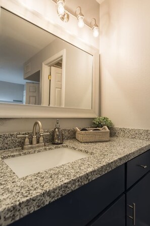 A bathroom with a granite countertop, a sink, a mirror with lights, a plant, and a door.
