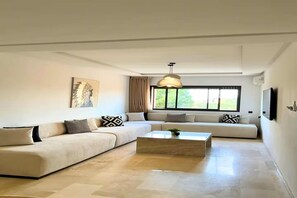 A modern living room with a sectional sofa, a coffee table, and a large window with curtains. - Luxury 3BR Residence - Gueliz - Pool-Walk to Majorelle (Marrakech)
