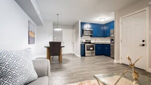 Een moderne woonkamer met een grijze bank, een eettafel met een stoel, een glazen salontafel, en een keuken met blauwe kastjes en roestvrijstalen apparatuur.