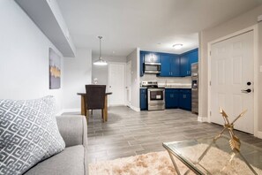 Una sala moderna con un sofá gris, una mesa de comedor con una silla, una mesa de centro de vidrio y una cocina con armarios azules y electrodomésticos de acero inoxidable.
