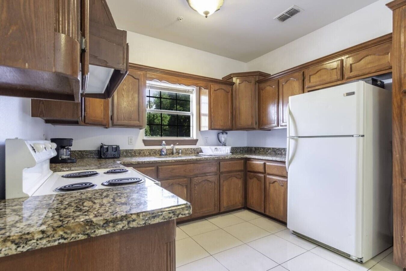 Una cocina con encimeras de granito, armarios de madera, un refrigerador blanco y una ventana con vista a follaje.