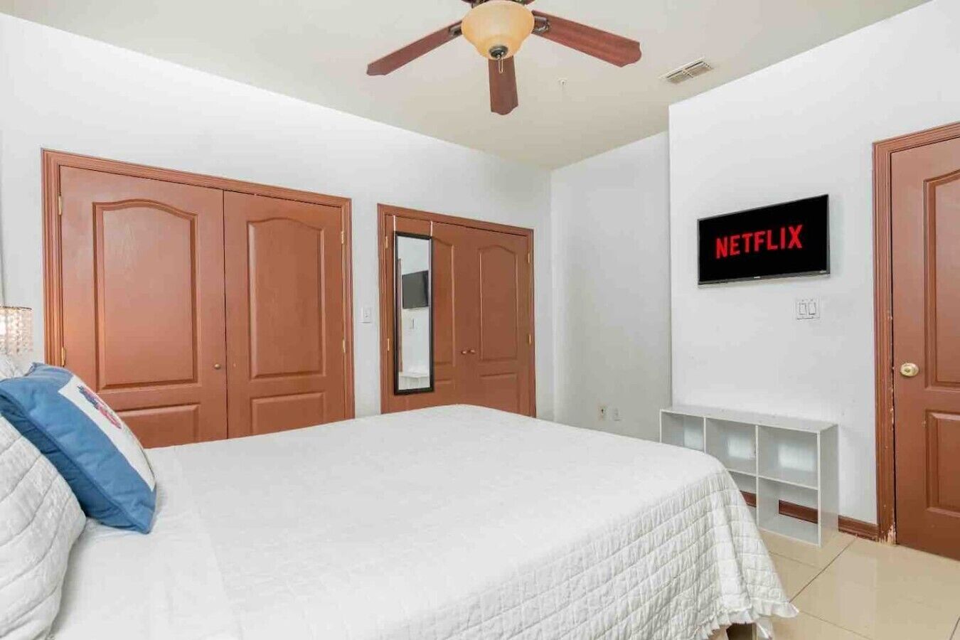 A bedroom with a bed, wooden wardrobe, a ceiling fan, a TV mounted on the wall, and a mirror.