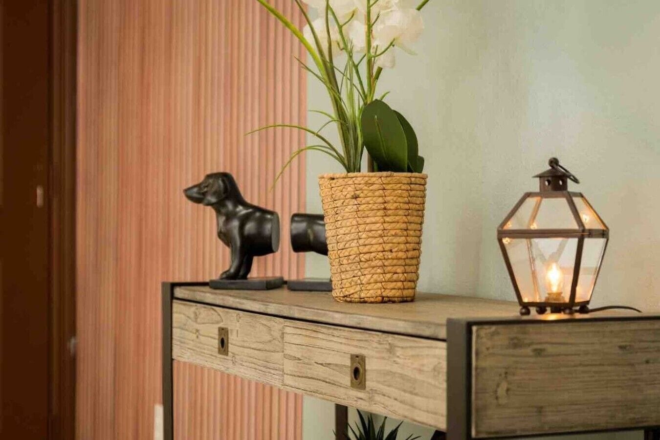 A wooden console table with a woven basket, a dog figurine, a potted plant, and a lit lantern.