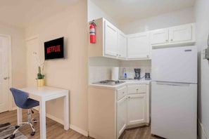 A small kitchenette with white cabinets, a stove, a sink, and a refrigerator. There is a small table with a chair and a potted plant. A fire extinguisher is mounted on the wall above the cabinets.