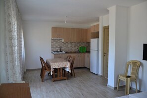 A compact kitchen with wooden cabinets, a dining table with chairs, a refrigerator, and a single chair in the corner.