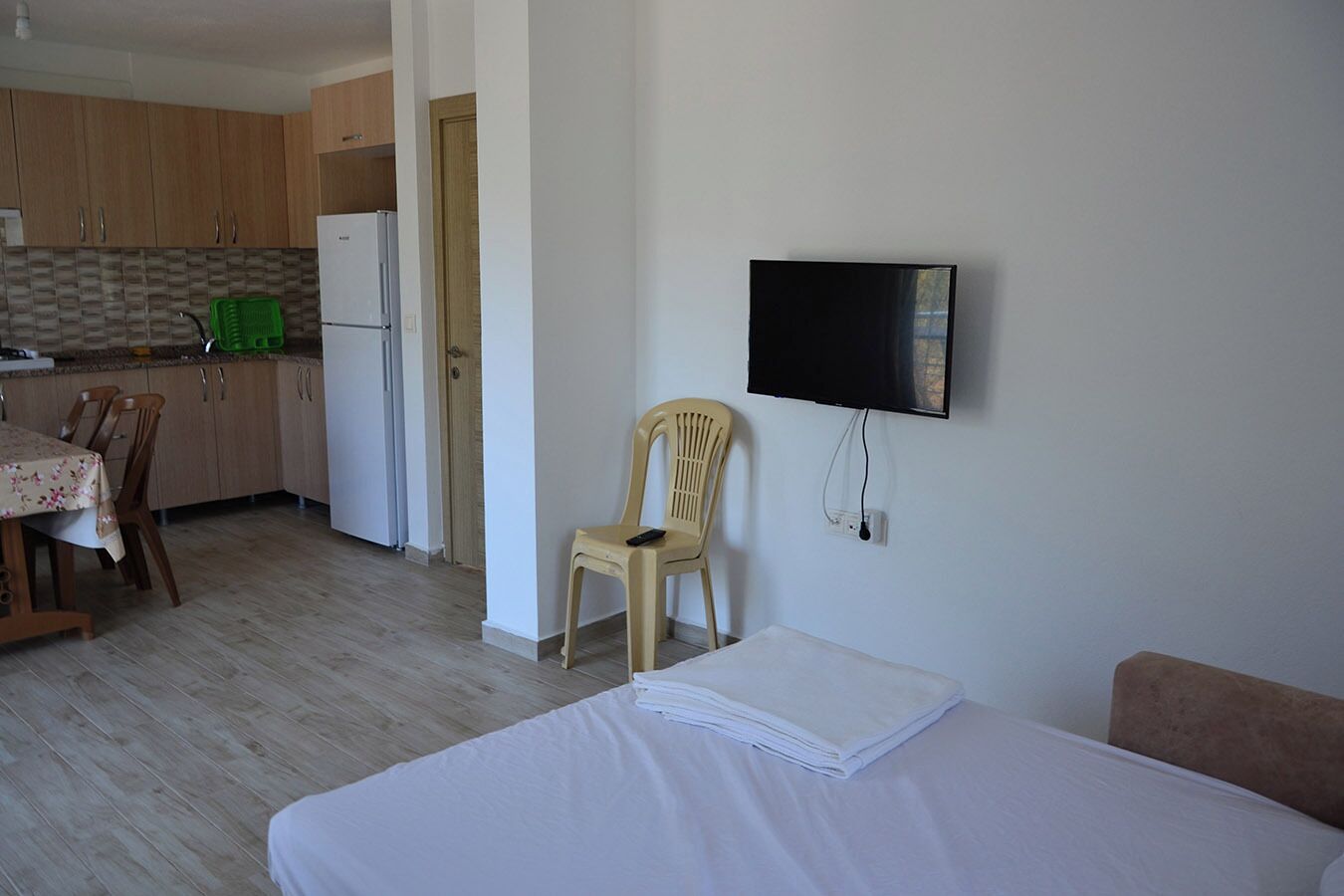 Quarto com cama, uma cadeira, televisão presa na parede, espaço de cozinha com armários e geladeira, e mesa de jantar com cadeiras.