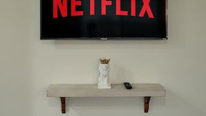 Ein an der Wand befestigter Fernseher, auf dem in roten Buchstaben 'NETFLIX' steht. Darunter eine weiße Statue mit goldener Krone auf einem hölzernen Regal.