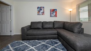 Ein Wohnzimmer mit einem grauen Ecksofa, einem blauen Teppich mit Muster und zwei Wandbildern.