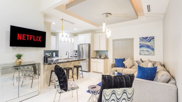 Uma sala de estar moderna com um sofá, uma área de jantar com mesa de vidro e uma cozinha ao fundo.
