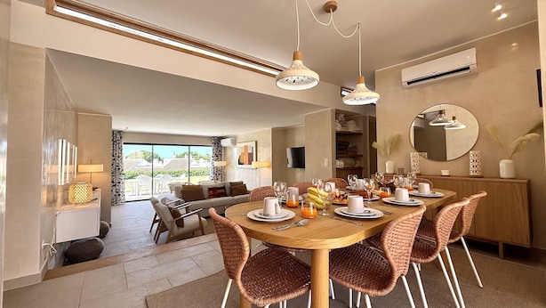 A dining area with a wooden table set for six, surrounded by wicker chairs, with a view of the outdoors through large windows. - Vilamoura Golf Villa 4 by Homing (Loulé)
