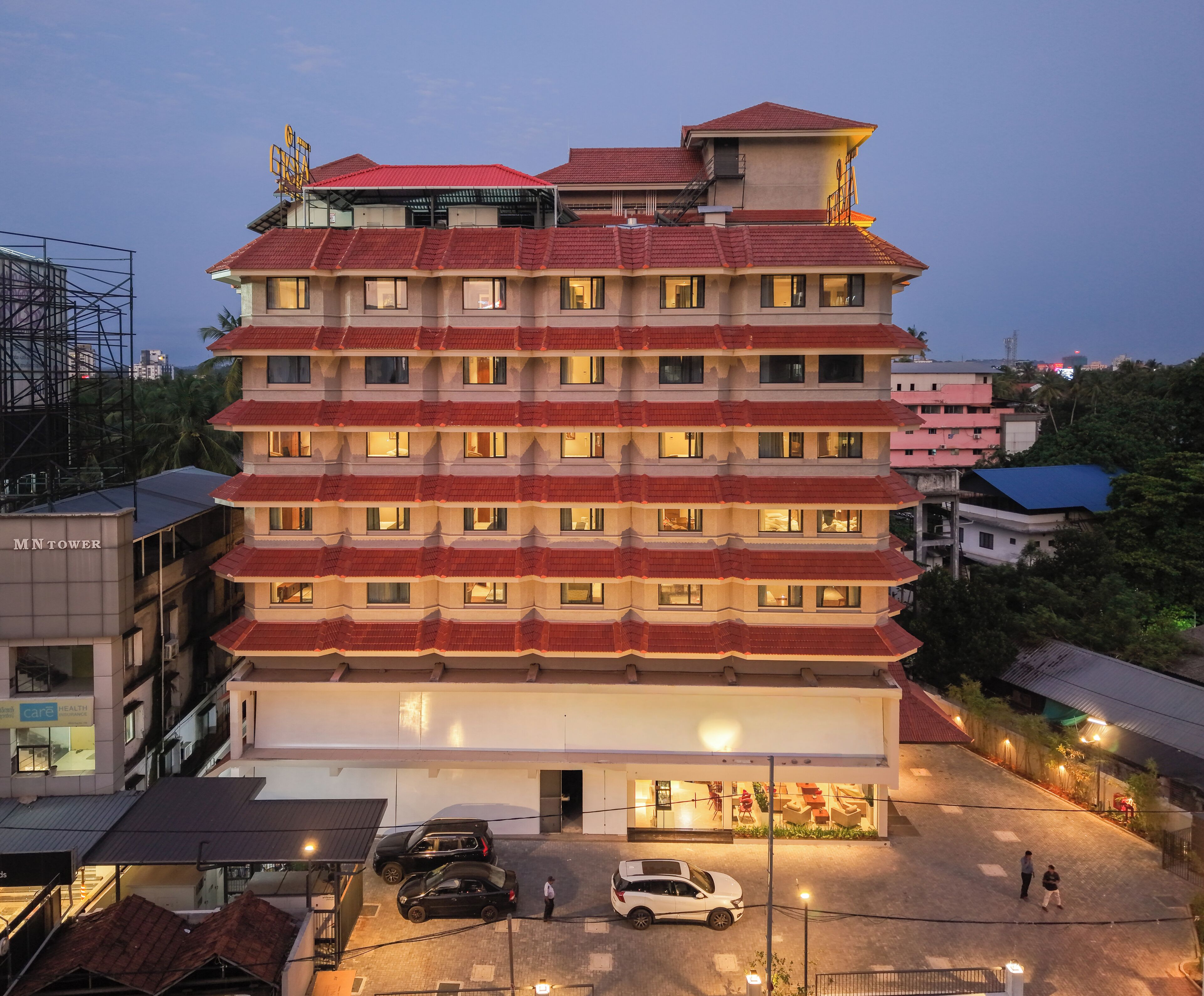 A multi-story hotel with a red-tiled roof, multiple balconies, and a ground-level entrance with a car parking area.