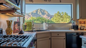 A kitchen with a stove, sink, and dishwasher, a large window with a mountain view, and decorative wall elements. - Luxury Sedona Views, Spa, Sauna, Cold Plunge (Sedona)