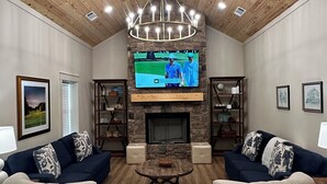 Ein geräumiges Wohnzimmer mit Holzboden, einem Steinkamin, zwei Sofas, einem Couchtisch und einem Fernseher, der ein Golfspiel zeigt.