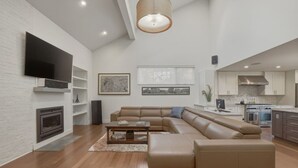 Ein modernes Wohnzimmer mit Ecksofa, Flachbildfernseher, Kamin und einer Küche im Hintergrund.