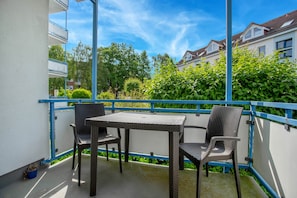 Un balcón con mobiliario de mimbre, baranda azul y vista a edificios residenciales y vegetación.