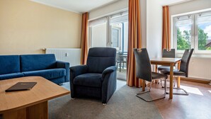 A room with a blue sofa, a dark armchair, a wooden table, and a dining area with chairs. - Apartment 'Ringstr. 21, 2. OG Links WE55' with Balcony and Wi-Fi (Binz)
