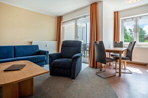 A room with a blue sofa, a dark armchair, a wooden table, and a dining area with chairs. - Apartment 'Ringstr. 21, 2. OG Links WE55' with Balcony and Wi-Fi (Binz)