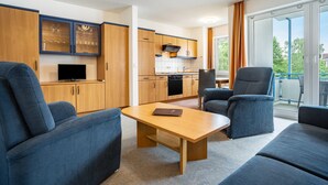 Ein modernes Wohnzimmer mit einer Couch, einem Tisch und einem Fernseher. Es gibt eine Küchenzeile mit Schränken und einem Fenster.