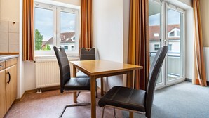 Ein Zimmer mit Holztisch, zwei schwarzen Stühlen, einem Fenster mit Vorhängen, einer Heizung und Blick auf Gebäude.