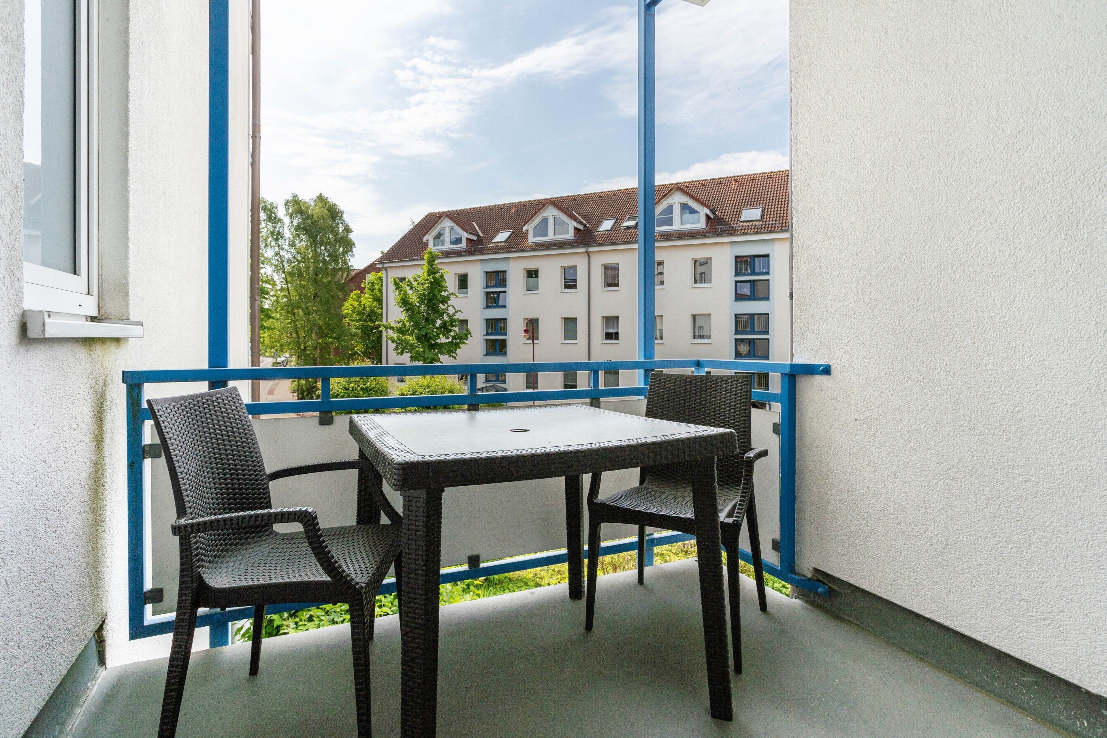Un balcon avec une table et des chaises, une vue sur un immeuble avec un toit en tuiles et une rambarde bleue.