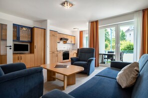Ein modernes Wohnzimmer mit Sofa, Sessel und Couchtisch, eine Küche mit Holzschränken und eine Glastür zum Außenbereich.