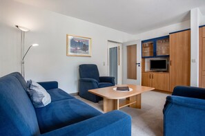 Un salon moderne avec un canapé, un fauteuil et une table sur laquelle se trouve un ordinateur portable. On aperçoit également une télévision et des meubles en bois à l’arrière-plan.