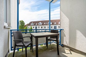 Un balcón con sillas de mimbre y una mesa, barandal azul, y vistas a una hilera de casas.