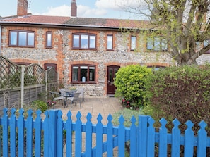 Une maison en pierre avec un toit recouvert de tuiles rouges, une clôture en bois peinte en bleu et une petite terrasse aménagée avec du mobilier d’extérieur.