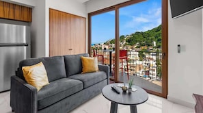 A modern living room with a grey sofa, a small round table, and a large sliding glass door to a balcony. - Rooftop Pool & Ocean Views in Zona Romántica (Puerto Vallarta)