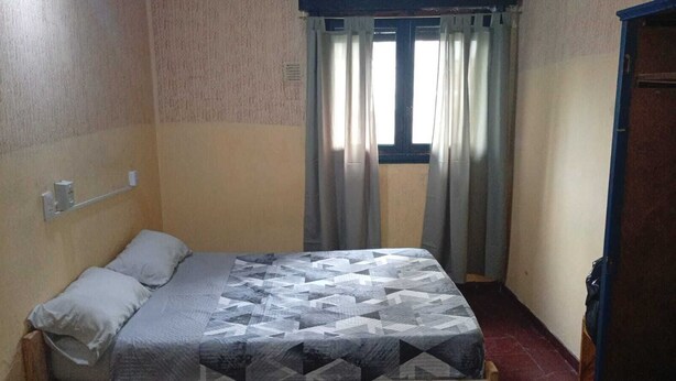 A small, single-bed room with a patterned bedspread, a window with curtains, and a door on the right. - Punto 11 (Buenos Aires)