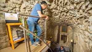 Un homme se tient sur une échelle en métal dans une pièce aux murs de pierre, où se trouvent un lit et une petite fenêtre.
