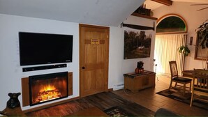 Een woonkamer met een open haard, een aan de muur bevestigde televisie, een houten deur en een eethoek met een tafel en stoelen.