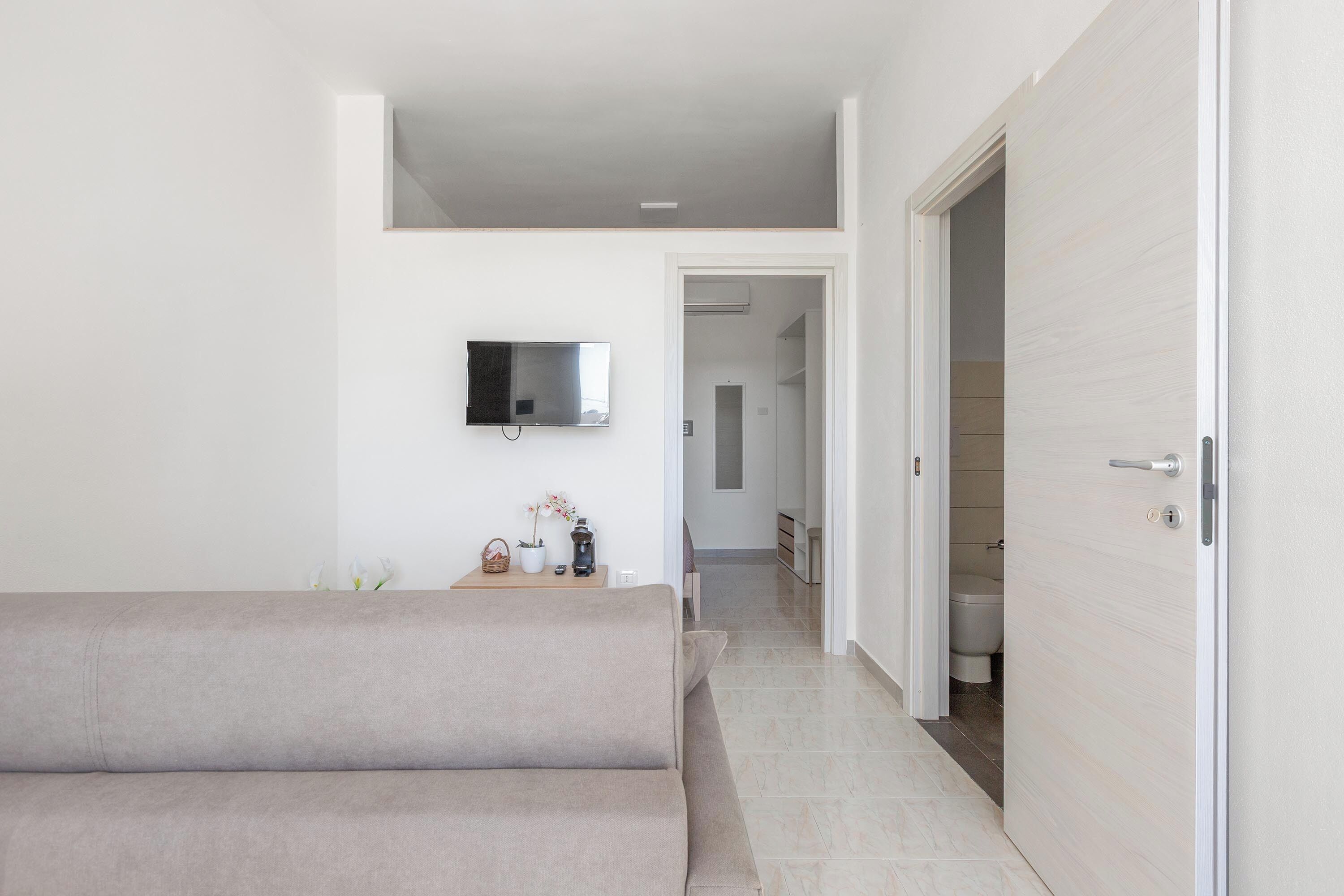 A modern living room with a sofa, a flat-screen TV mounted on the wall, a small table with a plant, and a door leading to a bathroom.