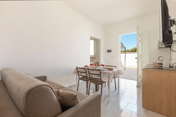 A living room with a sofa, a dining table with chairs, and a kitchen area with a counter and a television. - Borgo Climmena - Bilo con Patio (Manduria)