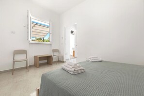 A minimalist hotel room with a bed, two chairs, a bench, a window with a view, and a door with a fan. - Borgo Climmena - Bilo con Patio (Manduria)