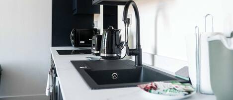 Una cocina moderna con encimera blanca, armarios negros, un hervidor y una cafetera.