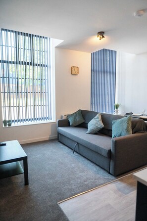 A modern living room with a grey sofa, a small table, and a window with vertical blinds. - Modern 1 Bedroom Apartment in Derby with Parking (Derby)