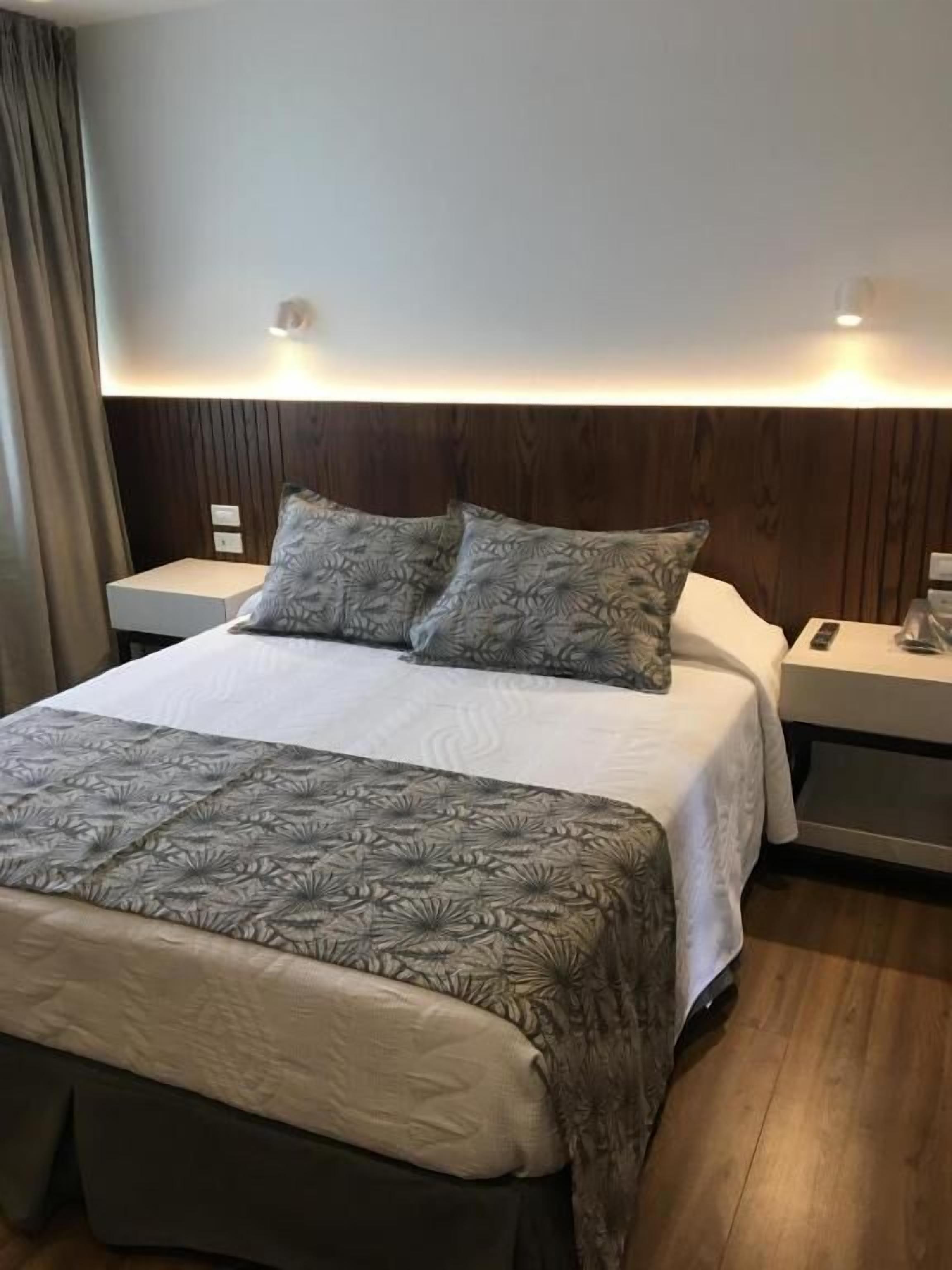 Une chambre d’hôtel avec un lit, deux tables de chevet, une petite table, un plancher en bois et un mur muni d’un luminaire.