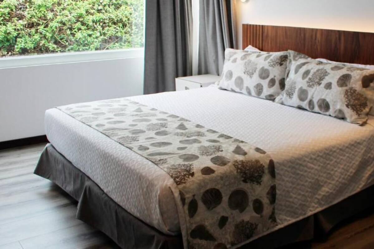 A neatly made bed with a patterned bedspread and pillows, a bedside table with a phone, and a window with curtains.
