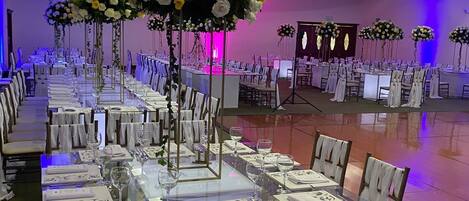 A banquet hall with long tables set for dining, chairs with white covers, and floral centerpieces.