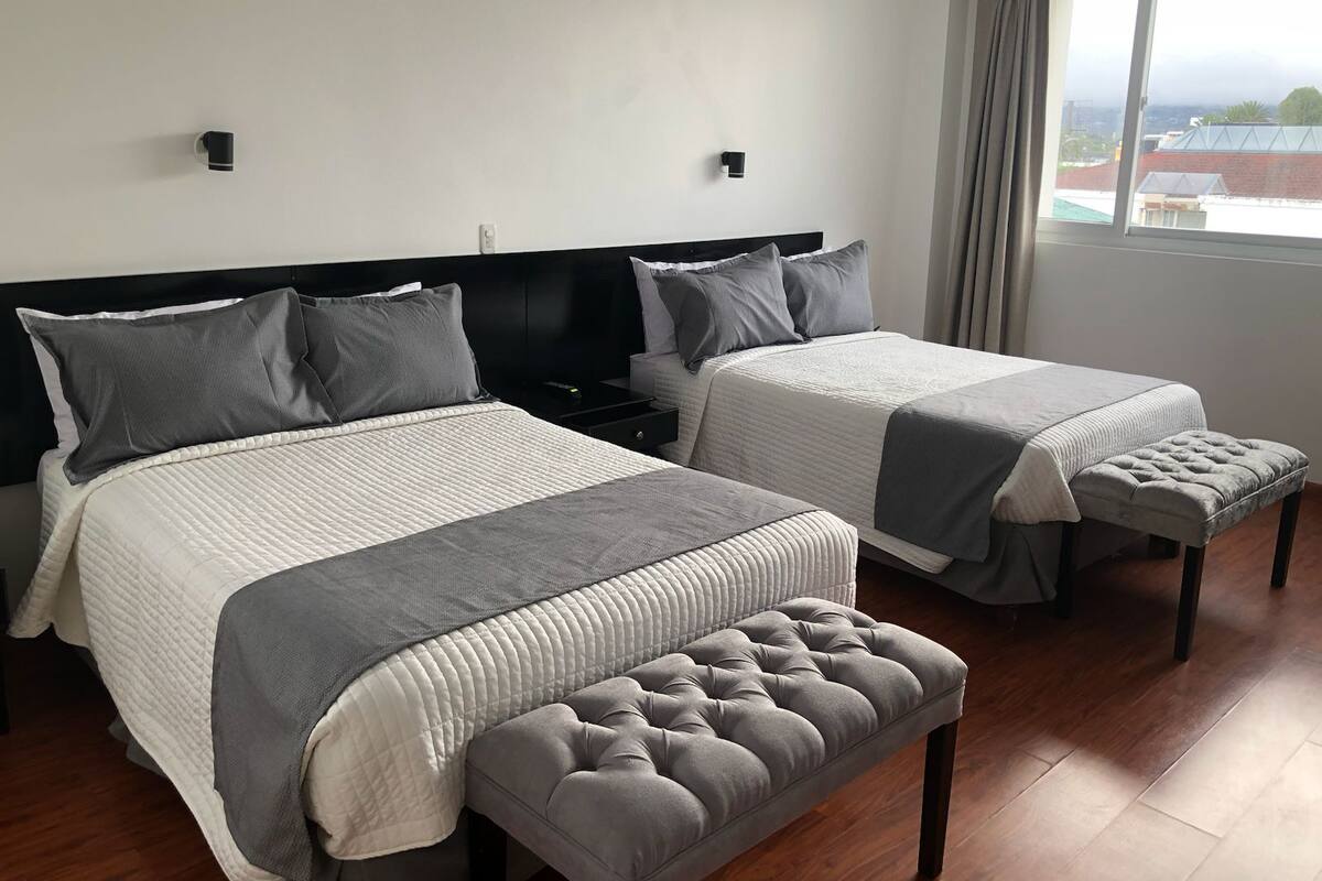 A double bed with a grey quilt, a bedside table, a grey bench, and a window with curtains.